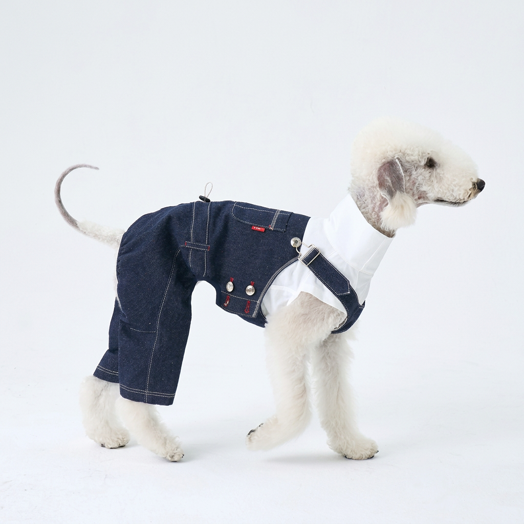 Side view of a white dog wearing dark indigo denim overalls with silver buttons, white stitching and rear-leg coverage over a separate white top.