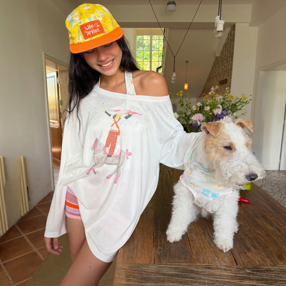 Model wearing the yellow Citrus Explorer human cap beside a white terrier on a wooden table; the dog is not wearing a hat.
